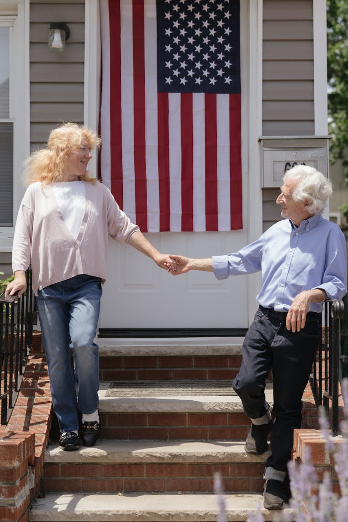 A happy senior couple holding hands on a porch with an American flag, symbolizing freedom and celebration.